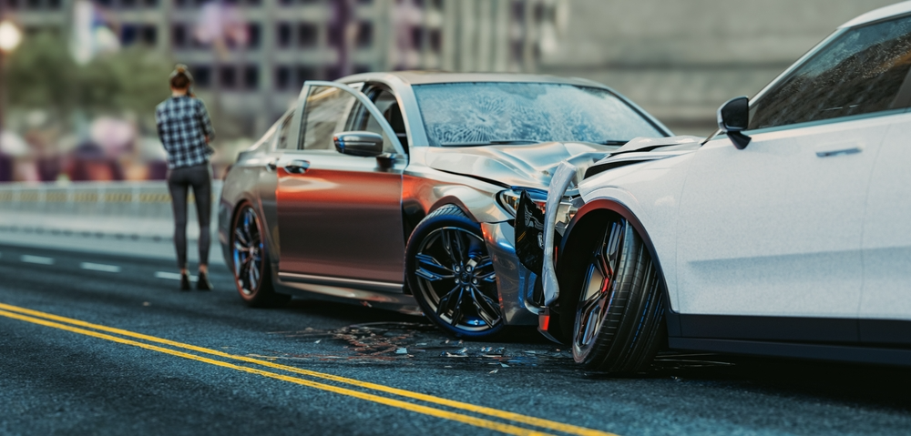 Two cars in a head-on collision on a city street. A person stands beside the road, inspecting the damage. The scene conveys shock and urgency.