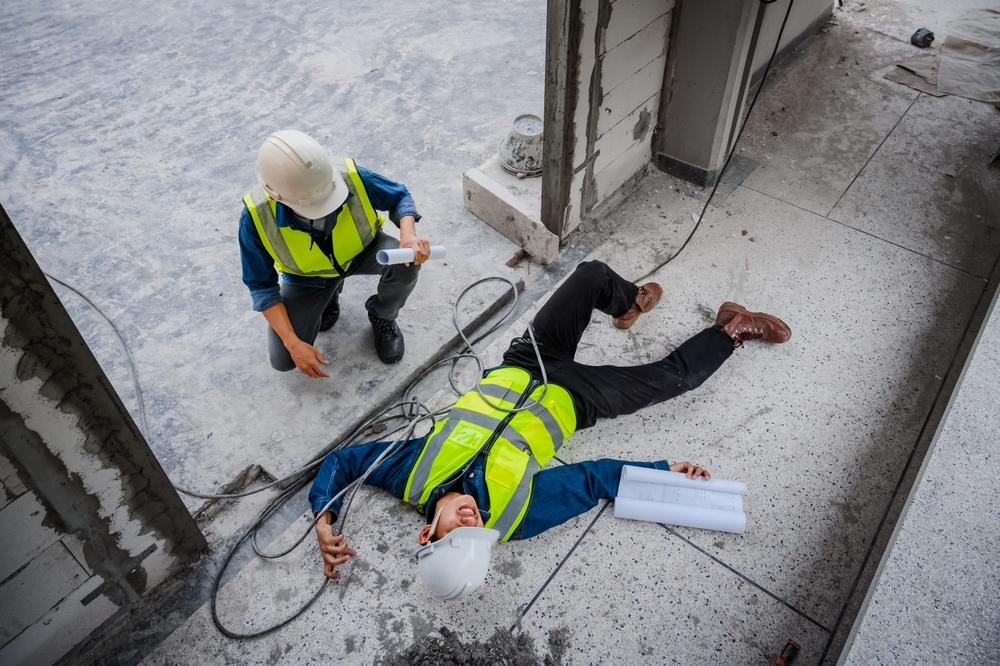 Two construction workers in safety gear; one is on the ground appearing hurt while the other kneels beside him, looking concerned. Tense and urgent setting.