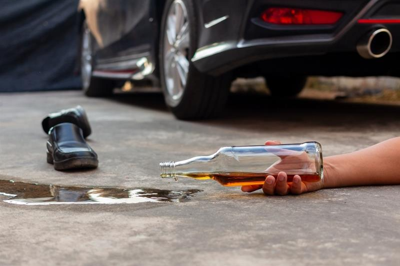 A person's hand holds an empty bottle, lying on a pavement beside a black car.