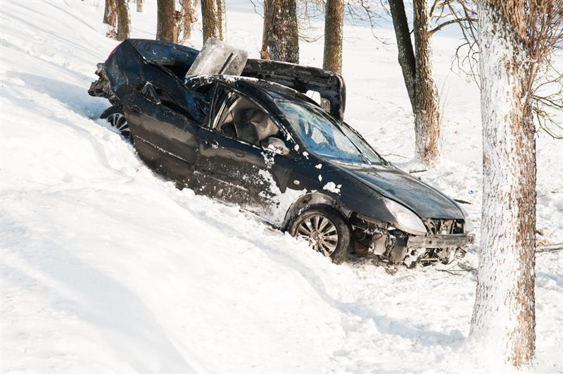 A black car is crashed into a snowy embankment amid bare trees, showing severe damage