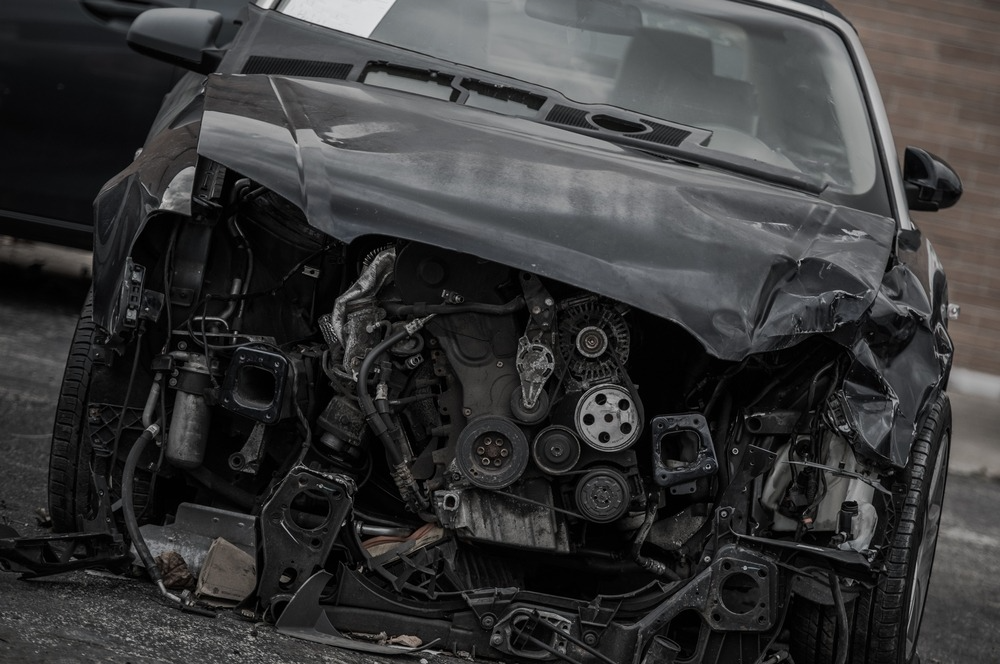 Front view of a heavily damaged black car with a crumpled hood, exposing its engine and mechanical components