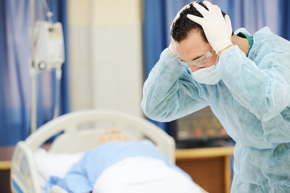 A medical professional in a protective gown and gloves holds his head in distress beside a hospital bed. The scene conveys tension and concern.
