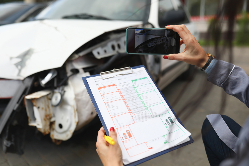 Person takes a photo of car damage with a smartphone, holding a clipboard with an accident report. The scene conveys documentation and assessment.