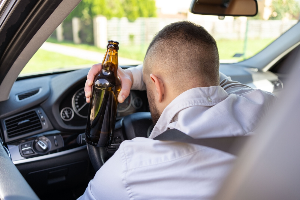 A person in a car leans on the steering wheel, holding an empty beer bottle, suggesting irresponsibility and potential danger.