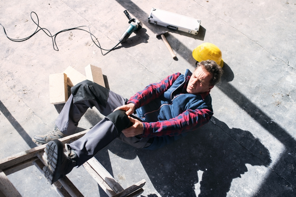 A construction worker sitting on the floor, holding his injured knee, appears in pain. Nearby are a hard hat, tools, and a fallen ladder.
